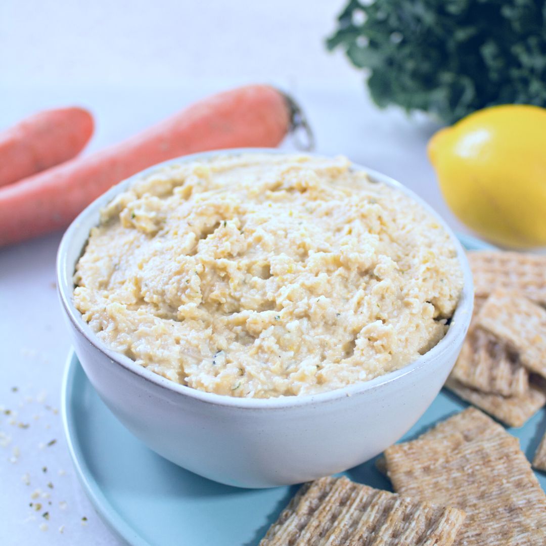 white bowl filled with lemon garlic hummus on blue plate with triscuits on the side and carrots and lemon and greens in the background