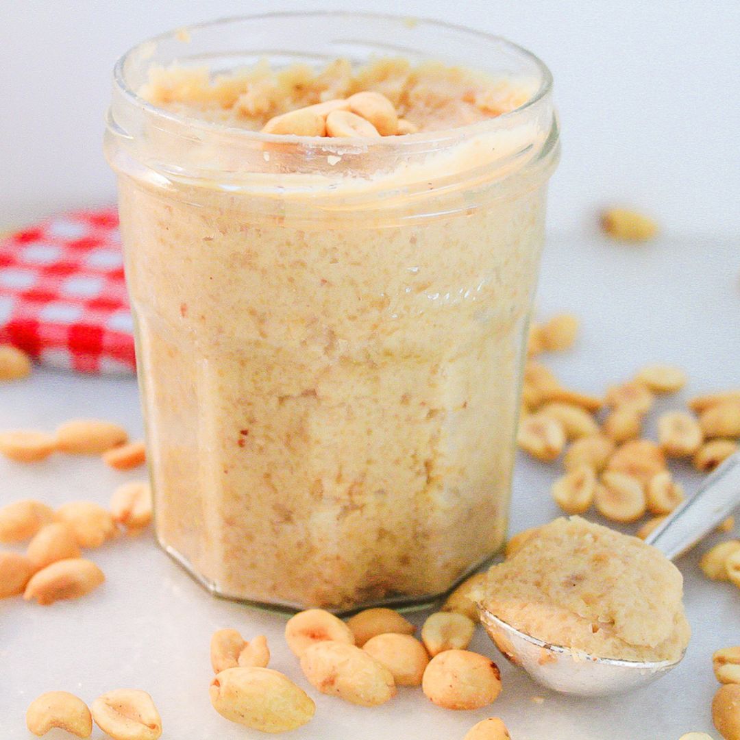 peanut butter coconut spread in a glass jar with a spoonful of it next to it and peanuts on the ground around it