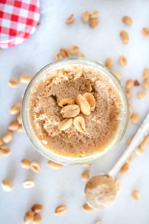peanut butter coconut spread in a glass jar with a spoonful of it next to it and peanuts on the ground around it