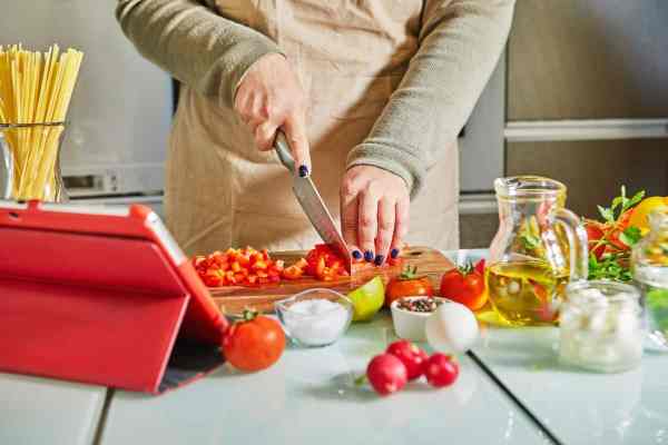 woman chopping tomatoes with ipad open next to her and ingredients around her